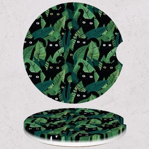 May include: Two car coasters with a cat and leaf design. The coasters feature a pattern of black cats peeking out from green leaves. One coaster is round with a cutout for a car cup holder, and the other is a full circle.