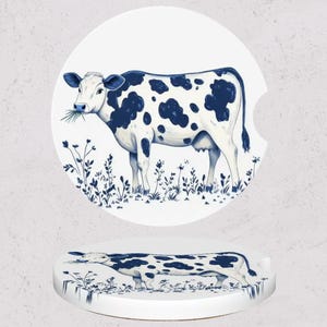 May include: Two white ceramic car coasters with a blue and white cow design. The cow is standing in a field of blue flowers and grass. The coasters are round with a small indentation for easy removal.