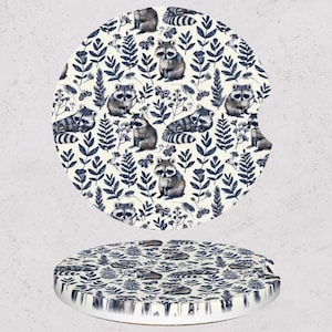 May include: Two round car coasters with a cream background. The coasters feature a repeating pattern of raccoons, leaves, and flowers in shades of blue and gray. One coaster has a cutout for a cup holder.