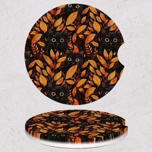 May include: Two round car coasters with a black cat and autumn leaf design. The coasters feature a black background with orange and yellow leaves and black cat faces. One coaster has a cutout for a car cup holder.