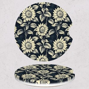 May include: Two car coasters with a sunflower design. The coasters are round with a dark blue background and a pattern of cream-colored sunflowers and leaves. One coaster has a notch for a car cup holder.