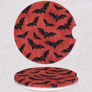 May include: Two round car coasters with a Halloween theme. The coasters feature a red background with a repeating pattern of black bats. One coaster has a cutout for a car cup holder. The coasters are made of a ceramic material.