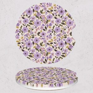 May include: Two round car coasters with a floral pattern. The design features purple flowers, green leaves, and small pink blossoms on a cream background. One coaster has a notch for easy removal from a car cup holder.