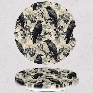 May include: Two car coasters with a raven and branch design. The coasters are round with a black and white raven and branch pattern on a cream background. One coaster has a cutout for a car cup holder.