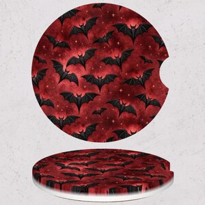 May include: Two round car coasters with a spooky bat design. The coasters feature a red background with a pattern of black bats and small white stars. One coaster has a curved cutout for easy placement in a car cup holder.