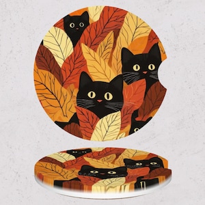 May include: Two round car coasters with a fall theme. The coasters feature black cats peeking out from behind autumn leaves in shades of yellow, orange, and brown. The coasters are made of a white absorbent material.