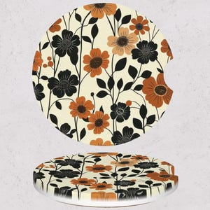 May include: Two round car coasters with a floral design. The coasters feature a cream background with black, orange, and brown flowers and leaves. One coaster has a cutout for a car cup holder.