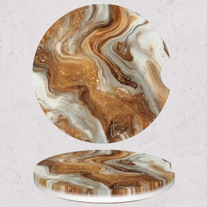 May include: Two car coasters with a swirling, abstract design in shades of gold, brown, and white. One coaster is shown flat, and the other is angled, revealing the side profile and a small notch for easy removal.