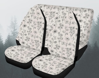 May include: Set of two car seat covers with a cream-colored background and a delicate black floral pattern. The covers have black trim and are designed to fit standard car seats. The floral design includes various flower and leaf shapes.