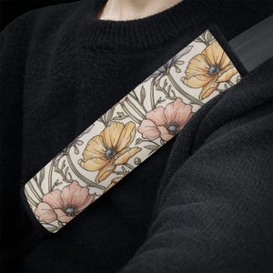 May include: A white car seatbelt cover with a floral pattern featuring yellow, pink, and green flowers. The flowers are stylised and have a vintage aesthetic.