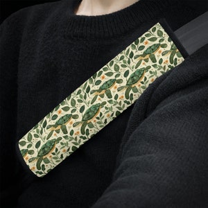May include: A beige car seatbelt cover with a repeating pattern of green sea turtles and yellow flowers on a cream background.