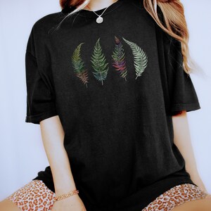 Puede incluir: Una camiseta negra de gran tama&ntilde;o con un gr&aacute;fico de cuatro hojas de helecho de colores. La camiseta la lleva puesta una persona con el pelo largo rubio y pantalones cortos de estampado de leopardo.