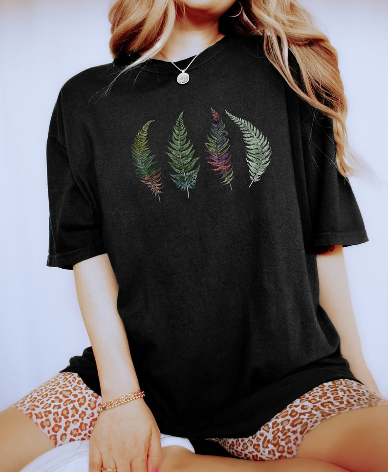 Puede incluir: Una camiseta negra de gran tama&ntilde;o con un gr&aacute;fico de cuatro hojas de helecho de colores. La camiseta la lleva puesta una persona con el pelo largo rubio y pantalones cortos de estampado de leopardo.