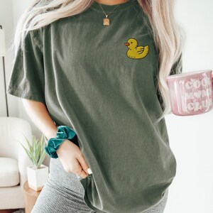May include: A green oversized t-shirt with a yellow rubber ducky graphic on the left chest. The shirt is being worn by a person with long blonde hair. The person is holding a pink mug with the text "Cool Mom Club" on it.
