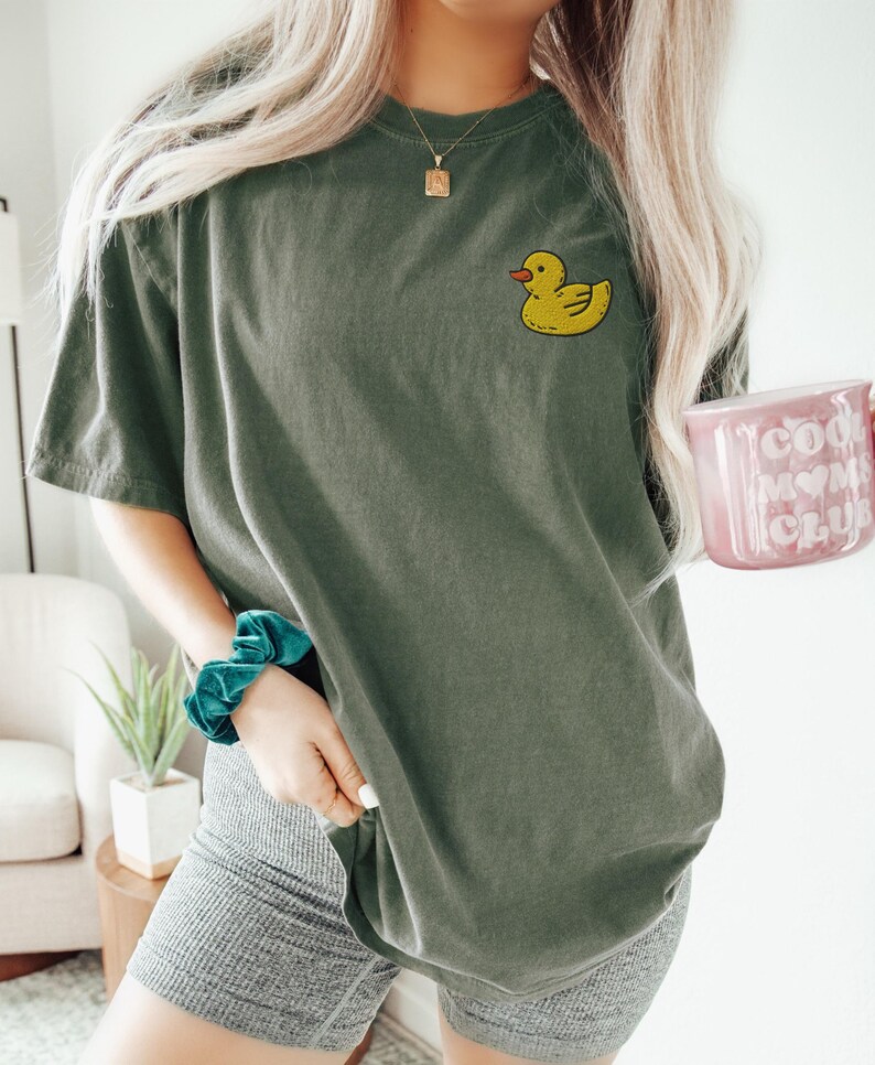 May include: A green oversized t-shirt with a yellow rubber ducky graphic on the left chest. The shirt is being worn by a person with long blonde hair. The person is holding a pink mug with the text "Cool Mom Club" on it.