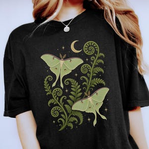 May include: Black t-shirt featuring a green moth design with fern-like foliage, a crescent moon, and stars. The text "ZEN & SHAPE" is printed at the bottom. The design is centered on the shirt.