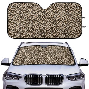 May include: A rectangular car windshield sun shade featuring a tan and black leopard print pattern with a black border. The sun shade is shown both as a standalone item and installed inside the front windshield of a white vehicle.