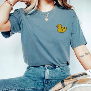 May include: A blue t-shirt with a yellow rubber ducky graphic on the front. The shirt is being worn by a person with a necklace and bracelets.