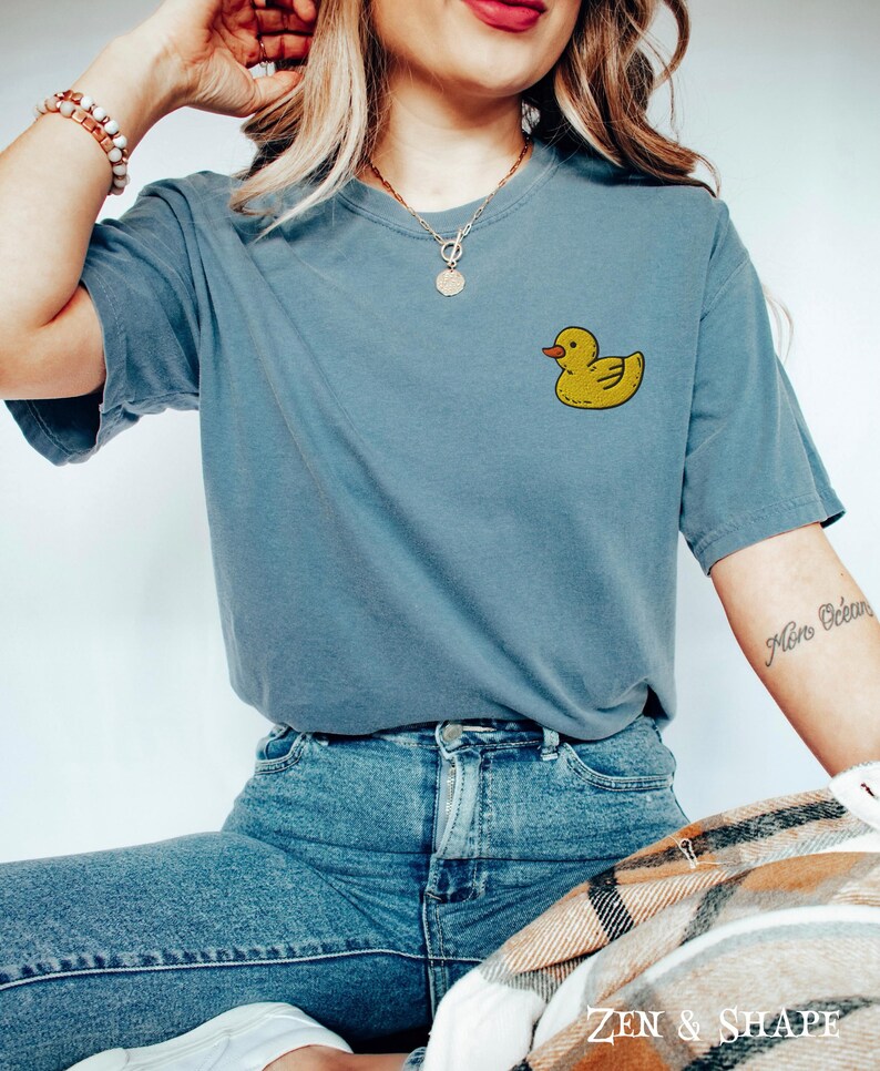 May include: A blue t-shirt with a yellow rubber ducky graphic on the front. The shirt is being worn by a person with a necklace and bracelets.