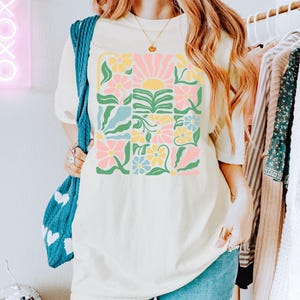 K&ouml;nnte beinhalten: Cremefarbenes T-Shirt mit einem farbenfrohen Blumendesign und einer Sonnengrafik. Das Design enth&auml;lt rosa, gelbe, blaue und gr&uuml;ne Elemente. Der Text "ZEN & SHAPE" ist unten rechts auf dem Shirt aufgedruckt.