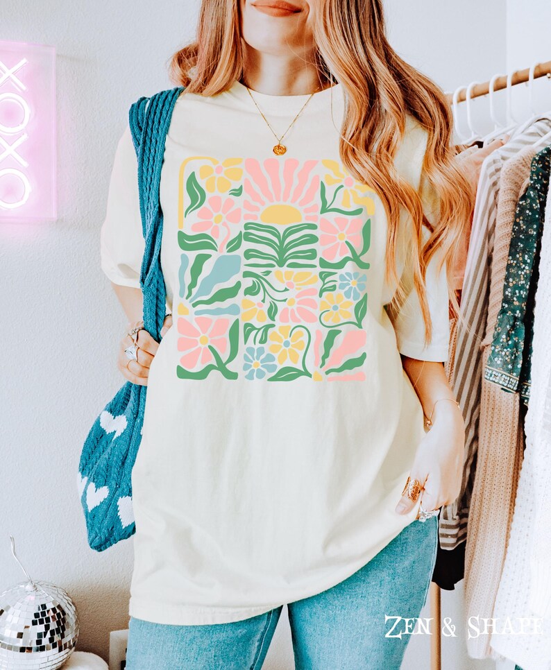 K&ouml;nnte beinhalten: Cremefarbenes T-Shirt mit einem farbenfrohen Blumendesign und einer Sonnengrafik. Das Design enth&auml;lt rosa, gelbe, blaue und gr&uuml;ne Elemente. Der Text "ZEN & SHAPE" ist unten rechts auf dem Shirt aufgedruckt.
