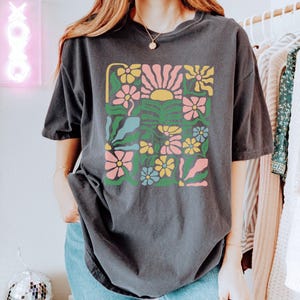 K&ouml;nnte beinhalten: Dunkelgraues T-Shirt mit einem farbenfrohen Blumendesign und einer Sonne. Das Design umfasst rosa, gelbe, blaue und gr&uuml;ne Blumen. Das Shirt hat eine lockere Passform und kurze &Auml;rmel. Der Text "ZEN & SHAPE" befindet sich unten rechts.