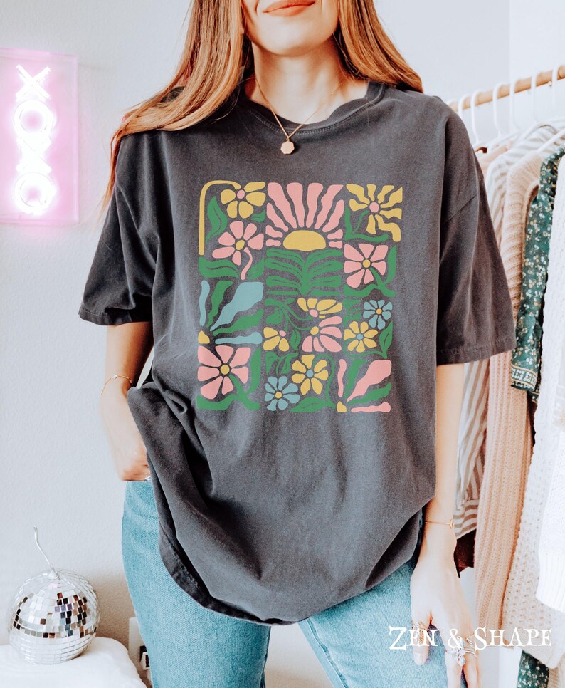 K&ouml;nnte beinhalten: Dunkelgraues T-Shirt mit einem farbenfrohen Blumendesign und einer Sonne. Das Design umfasst rosa, gelbe, blaue und gr&uuml;ne Blumen. Das Shirt hat eine lockere Passform und kurze &Auml;rmel. Der Text "ZEN & SHAPE" befindet sich unten rechts.