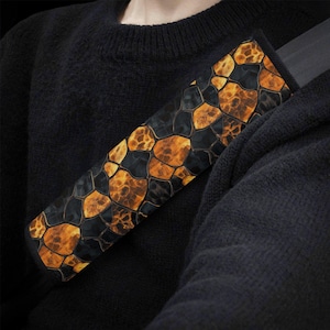 May include: A seat belt cover featuring a pattern of black and orange irregular shapes. The cover is rectangular and made of a soft material. The design resembles a mosaic or reptile skin.