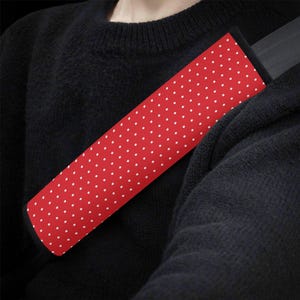 May include: A red seat belt cover with a white polka dot pattern. The cover is designed to fit over a standard seat belt, providing a decorative and potentially more comfortable experience. The cover is shown against a black background.