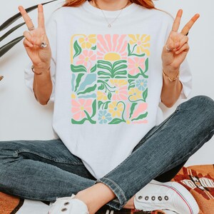 K&ouml;nnte beinhalten: Wei&szlig;es T-Shirt mit einem farbenfrohen Blumendesign in einem Rastermuster. Das Design umfasst rosa, gelbe und blaue Blumen, gr&uuml;ne Bl&auml;tter und eine Sonne. Der Text "ZEN & SHAPE" ist unten sichtbar.