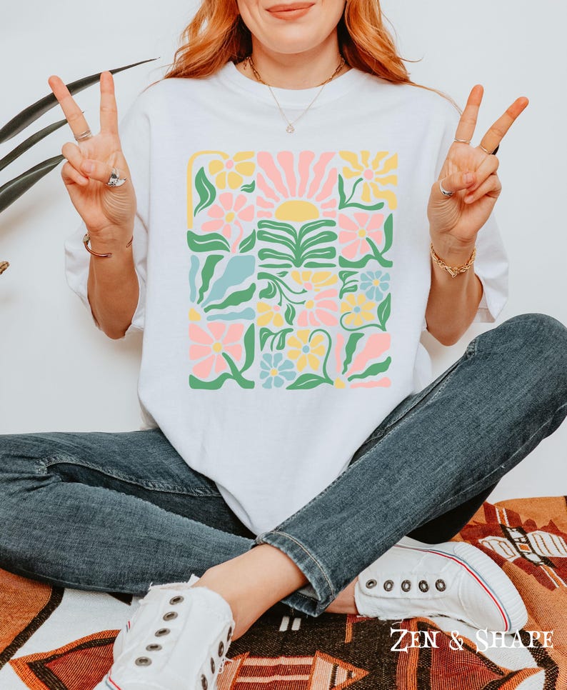 K&ouml;nnte beinhalten: Wei&szlig;es T-Shirt mit einem farbenfrohen Blumendesign in einem Rastermuster. Das Design umfasst rosa, gelbe und blaue Blumen, gr&uuml;ne Bl&auml;tter und eine Sonne. Der Text "ZEN & SHAPE" ist unten sichtbar.