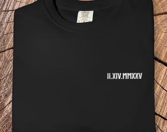 May include: Black crew neck t-shirt with white embroidered Roman numerals "II.XIV.MMXXV" on the front. The shirt is folded on a wooden surface. The brand name "ZEN & SHAPE" is printed at the bottom.