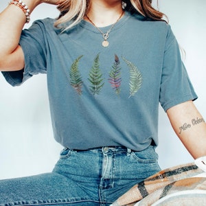 Puede incluir: Una camiseta azul con un gr&aacute;fico de cuatro hojas de helecho en tonos de verde, morado y rojo. La camiseta la lleva puesta una persona con el pelo largo y rubio.