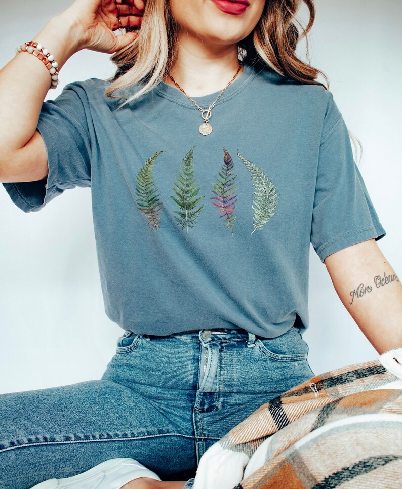Puede incluir: Una camiseta azul con un gr&aacute;fico de cuatro hojas de helecho en tonos de verde, morado y rojo. La camiseta la lleva puesta una persona con el pelo largo y rubio.