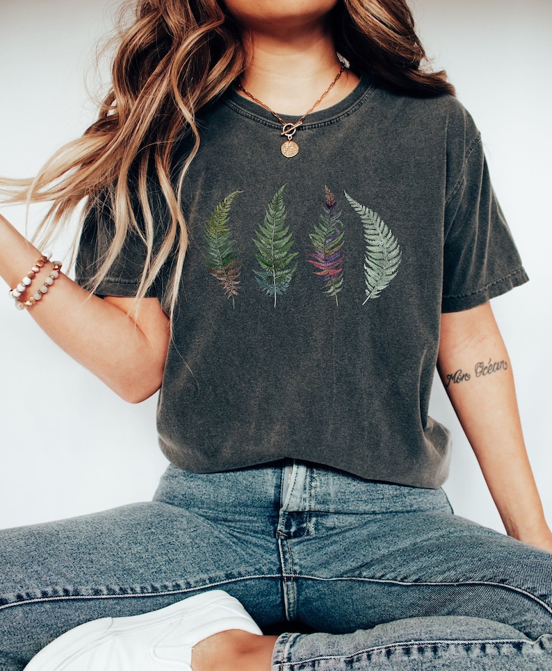 Puede incluir: Una camiseta gris oscuro con un gr&aacute;fico de cuatro helechos verdes. Los helechos est&aacute;n dispuestos en una fila y tienen un aspecto ligeramente descolorido. La camiseta tiene un corte holgado y est&aacute; hecha de una tela suave y c&oacute;moda.