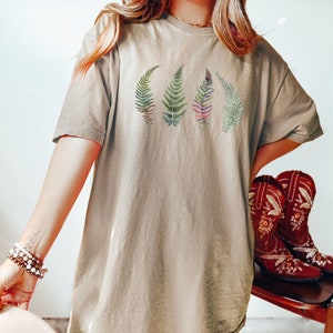 Puede incluir: Una camiseta de gran tama&ntilde;o de color marr&oacute;n claro con un gr&aacute;fico de cuatro hojas de helecho en tonos de verde, morado y rosa.