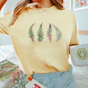 Puede incluir: Una camiseta amarilla de manga corta con un gr&aacute;fico de cuatro hojas de helecho en tonos de verde, morado y rosa. La camiseta la lleva puesta una persona con el pelo rubio y se combina con unos pantalones cortos de color azul claro.
