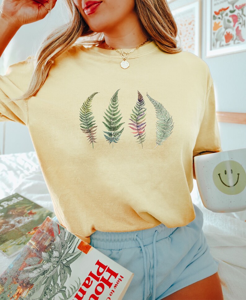 Puede incluir: Una camiseta amarilla de manga corta con un gr&aacute;fico de cuatro hojas de helecho en tonos de verde, morado y rosa. La camiseta la lleva puesta una persona con el pelo rubio y se combina con unos pantalones cortos de color azul claro.