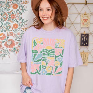K&ouml;nnte beinhalten: Ein helllila T-Shirt mit einem farbenfrohen Blumenmuster. Das Design zeigt Blumen, eine Sonne und Bl&auml;tter in Rosa, Gelb und Blau. Der Text "ZEN & SHAPE" ist unten auf dem Shirt aufgedruckt.
