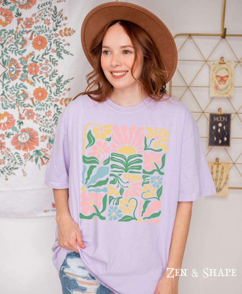 K&ouml;nnte beinhalten: Ein helllila T-Shirt mit einem farbenfrohen Blumenmuster. Das Design zeigt Blumen, eine Sonne und Bl&auml;tter in Rosa, Gelb und Blau. Der Text "ZEN & SHAPE" ist unten auf dem Shirt aufgedruckt.