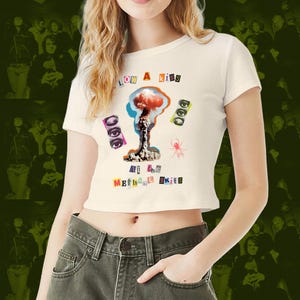 Könnte beinhalten: Cremefarbenes Crop-Top mit einem grafischen Design mit einer Atompilzwolke, Augen und einer Spinne. Der Text auf dem Shirt lautet "LOW A KISS" und "At the Methane Kits". Das Shirt hat kurze Ärmel und einen runden Ausschnitt.