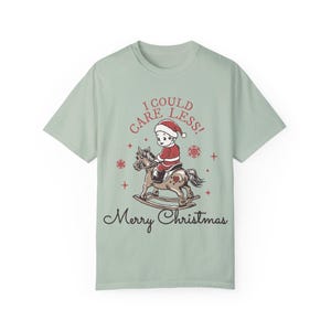Könnte beinhalten: Hellgrünes T-Shirt mit einem weihnachtlichen Design. Es zeigt eine Cartoon-Illustration eines Kindes im Weihnachtsmann-Kostüm auf einem Schaukelpferd, mit dem Text "I COULD CARE LESS!" und "Merry Christmas".