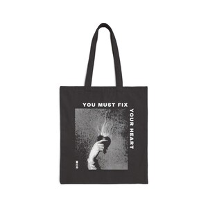 Könnte beinhalten: Schwarze Tragetasche mit weißem Text "YOU MUST FIX YOUR HEART". Die Tasche zeigt eine Grafik einer Hand, die ein Herz hält, aus dem Rauch oder Flammen aufsteigen. Die Buchstaben "MCR" sind unten links aufgedruckt.