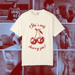 Puede incluir: Camiseta color crema con un gráfico de cerezas rojas y el texto "She's my cherry pie!". El gráfico presenta dos cerezas con el número "4" en cada una. Una prenda de vestir informal.