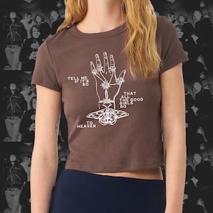 Könnte beinhalten: Ein braunes Crop-Top mit einer weißen Grafik einer Hand mit Schmuck und einer Motte. Der Text auf dem Shirt lautet "TELL ME IF IT'S SO", "THAT ALL THE GOOD GIRLS GO" und "TO HEAVEN".