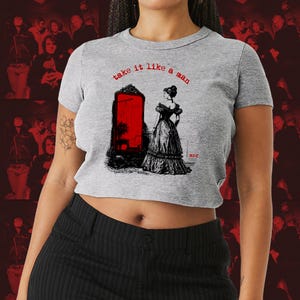 Könnte beinhalten: Graues Crop-Top mit dem roten Text "take it like a man". Das Shirt zeigt eine Schwarz-Weiß-Illustration einer Frau in einem langen Kleid vor einem roten Spiegel.