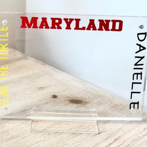 May include: Clear acrylic sign with the word "MARYLAND" in red block letters. The words "FEAR THE TURTLE" are in yellow vertically on the left, and "DANIELLE" in black vertically on the right. The sign is supported by a clear stand.