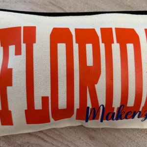 May include: Cream-colored canvas pencil case with the word "FLORIDA" in large orange block letters. Below, in a cursive navy blue font, is the name "Makenzie". The case has a black zipper.