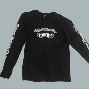 May include: Black long-sleeve shirt with the text "Original Mustache Club" and "OMC" with a mustache graphic in white. The sleeves have the text "Original Mustache Club" in white. The shirt is made of a soft material.