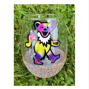 May include: A clear glass wine glass with a colorful tie-dye bear sticker on the front. The bottom of the glass is covered in iridescent glitter.